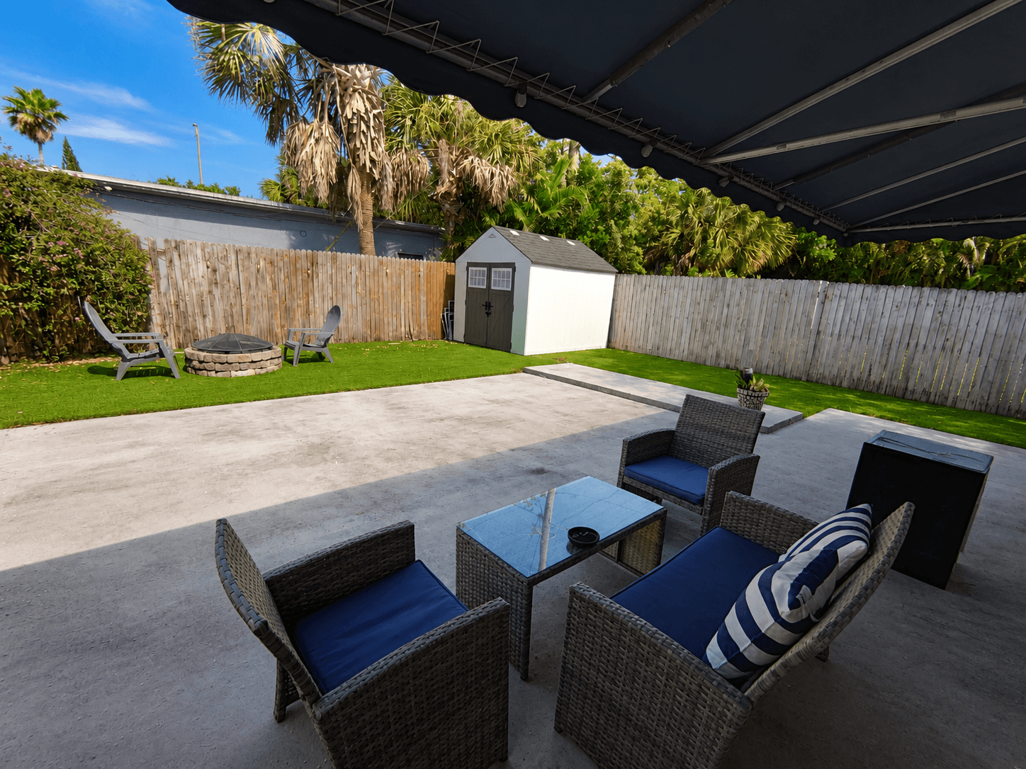 Fenced patio with wicker lounge seating, retractable awning, and stone fire pit